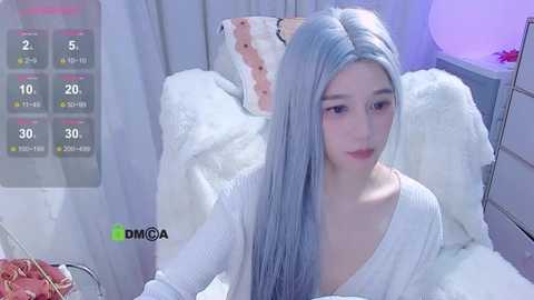 Media: Video of a young, light-skinned woman with long, pastel blue hair, wearing a white, off-shoulder sweater, sitting on a white, fluffy blanket. Background includes a pink pillow and a white dresser.