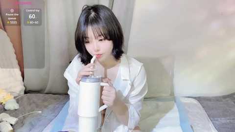 Media: Video of an East Asian woman with short black hair, in a white robe, drinking from a glass jar, indoors with a bed and curtains in the background.