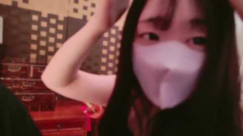 Media: A video of an Asian woman with long black hair, wearing a white surgical mask, standing in a dimly lit room with checkered wallpaper and a wooden staircase.