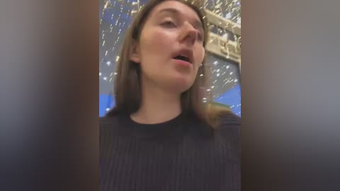 Media: Video of a woman with straight, medium-length brown hair, wearing a black ribbed sweater, singing with eyes closed, surrounded by a blurred background of golden lights and blue elements.