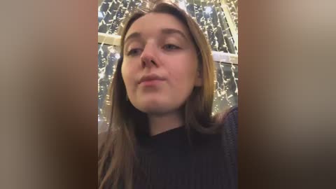 Media: A video of a young Caucasian woman with straight, shoulder-length light brown hair, wearing a dark sweater. She has a neutral expression and stands in front of a background with twinkling fairy lights, creating a warm, festive atmosphere.