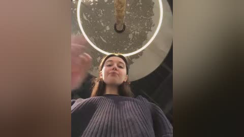 Media: A low-angle video of a young woman with long brown hair, wearing a dark blue sweater, standing under a circular, dimly lit ceiling fan. The background is blurred, with a muted color palette.