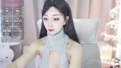 Media: Video of a young East Asian woman with long black hair, wearing a grey ribbed turtleneck sweater, sitting indoors with a blurred background.