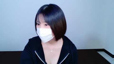 Media: Video of an East Asian woman with straight black hair, wearing a white surgical mask and black hoodie, indoors against a plain white background.