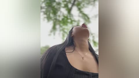 Media: Video of a woman with long, dark hair, wearing a black, low-cut top and necklace, looking upwards with eyes closed, set against a blurred, green, leafy background.