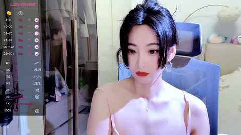 Media: Video of an Asian woman with pale skin, black hair, red lipstick, and a light-colored bra, sitting in a blue chair in a dimly lit room.