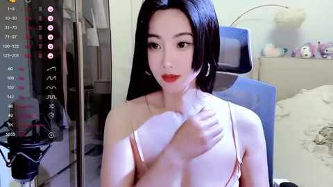 Media: A video of an East Asian woman with long black hair, fair skin, and red lipstick, wearing a light pink bra, in a modern bedroom.