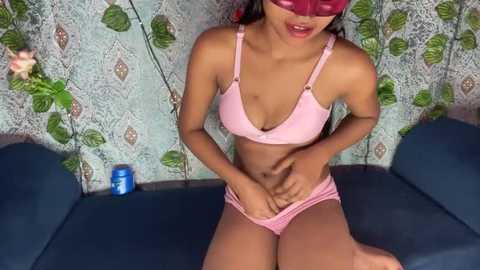 Media: Video of a petite, light-skinned woman with small breasts, wearing a pink bra and panties, sitting on a blue couch, blindfolded, against a patterned wallpaper background.