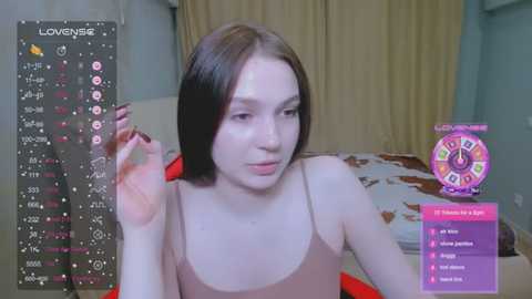 Media: Video of a young, pale-skinned woman with straight, dark brown hair, wearing a beige spaghetti-strap top, sitting on a bed in a room with light blue walls and beige curtains. She holds a red nail polish bottle.