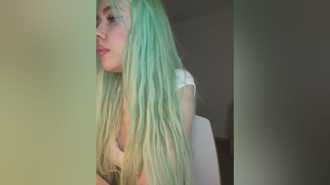 A video of a young woman with long, pastel green hair, fair skin, and a white top, gazing introspectively against a blurred background.