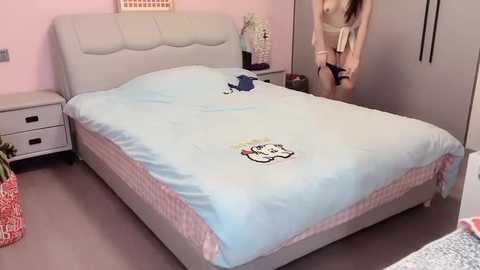 Media: Video of a minimalist, pink-walled bedroom with a beige bed, light blue duvet featuring cartoon characters, and a woman in a white robe standing by the closet.