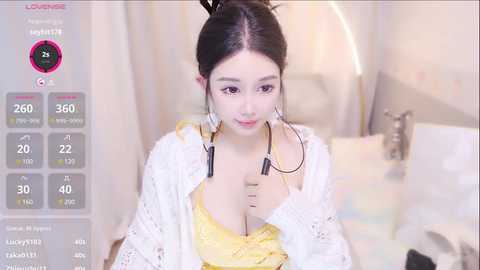 Media: Video of an Asian woman with fair skin and dark hair in a bun, wearing a yellow lace dress and a white crocheted cardigan, sitting in a pastel-colored room, holding a stethoscope.