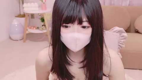 Media: Video of an East Asian woman with long black hair, wearing a white mask, in a soft, pastel-toned room with beige furniture and white walls.