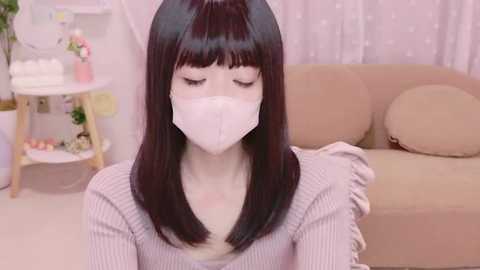 Media: Video of an Asian woman with straight, black hair and bangs, wearing a white face mask, grey sweater, and sitting indoors near a beige couch with a white pillow. Background includes a shelf with potted plants and white curtains.