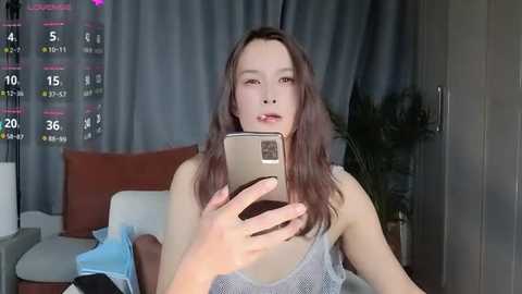 Media: A video of an Asian woman with long brown hair, wearing a light grey tank top, taking a selfie in a dimly lit room with grey curtains and a bed.