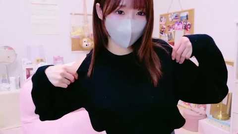 Media: Video of an Asian woman with straight, shoulder-length brown hair, wearing a black long-sleeved top, a face mask, and a pink shirt in the background. She stands in a light-colored room with white walls and a bulletin board.