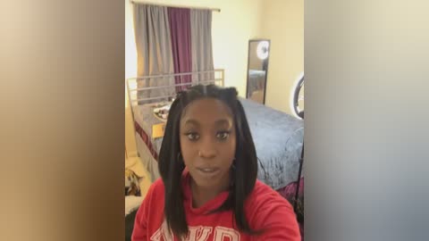 Media: Video of a young Black woman with braided hair in a red shirt, standing in a cozy bedroom with a bed, curtains, and a mirror.