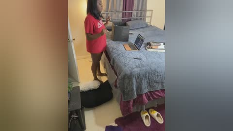 Media: Video of a pregnant Black woman in a red shirt, standing in a dimly-lit bedroom with a laptop, clothes, and a dog bed on the floor.