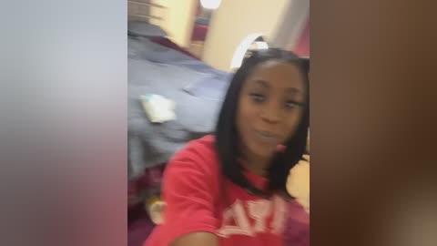 Media: A blurred video of a young Black woman with straight black hair, wearing a red T-shirt and a pink hair accessory. She's sitting on a bed with blue sheets in a dimly lit room.