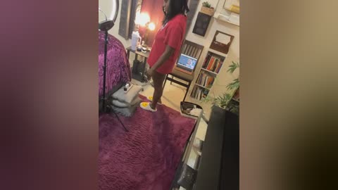 Media: Video of a woman in a red shirt and black pants, standing on a purple carpet in a cluttered room with a TV, bookshelves, and plants.