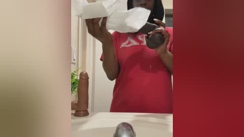 Media: A video shows a dark-skinned woman with short hair, wearing a red T-shirt, holding a tissue in her mouth, standing in a bathroom with a brown dildo on the counter.