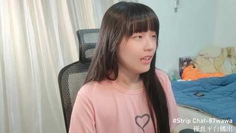 Media: Video of a young Asian woman with long black hair, wearing a pink T-shirt with a heart graphic, sitting on a black office chair, in a messy bedroom with blue bedding and orange clothes.