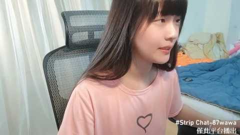 Media: Video of a young Asian girl with long black hair and a pink T-shirt with a heart design, sitting in a black mesh chair in a messy bedroom with blue bedding and white curtains.