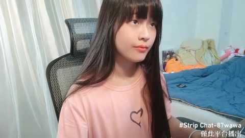 Media: A young Asian woman with long black hair and fair skin, wearing a pink T-shirt with a heart design, sits on a black mesh chair in a messy bedroom with a blue quilt and scattered clothes.