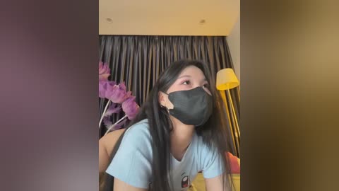 Media: Video of an Asian woman with long black hair, wearing a light blue t-shirt, black mask, and purple floral earrings, standing indoors near a curtain with a yellow lamp.