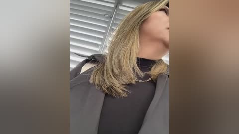 Media: A video of a woman with light skin, shoulder-length blonde hair, and a black turtleneck sweater. She stands in front of closed white blinds.