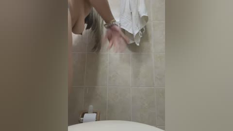 Media: Video of a nude woman with light skin and long blonde hair, seen from behind, removing a white towel in a beige-tiled bathroom.