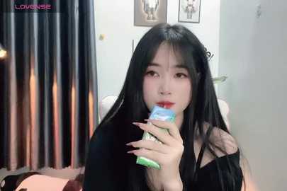 Media: Video of an East Asian woman with long black hair and fair skin, wearing a black off-shoulder top, drinking from a green and blue bottle, in a modern, minimally decorated room with framed art on the wall.