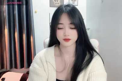 Media: Video of an Asian woman with long black hair, fair skin, and red lipstick, wearing a cream sweater, seated indoors against a white wall with a framed picture and curtains.