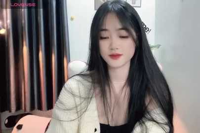 Media: Video of an East Asian woman with long black hair, fair skin, and a slender physique, wearing a white textured cardigan and black top, sitting indoors with a soft, warm light.