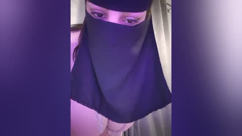 Media: Video of a woman with light skin, wearing a black niqab that covers her face except for her eyes, standing indoors in front of a purple backdrop.