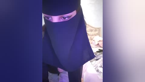 Media: A video of a woman wearing a black niqab with her face partially visible, standing in a cluttered bedroom with a bed and various objects in the background.