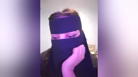 Media: Video of a woman in black niqab, face partially covered, with light skin, brown hair, and a dark scarf. Background is blurred with purple and white tones.