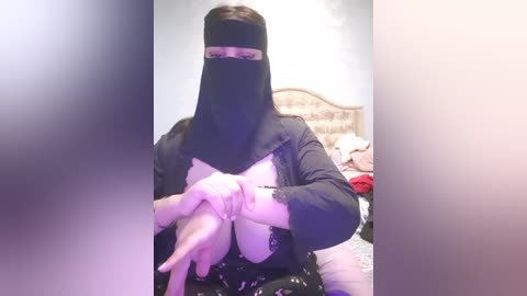 Media: Video of a person wearing a black niqab, pink gloves, and a white shirt, standing in a bedroom with a bed and scattered clothes in the background.