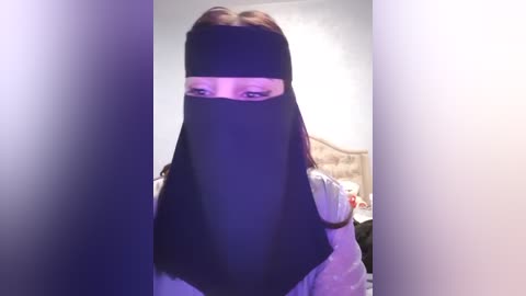 Media: Video of a person wearing a black face veil, with light skin, brown hair, and white shirt. The background shows a dimly lit, blurred bedroom with a bed and pillows.