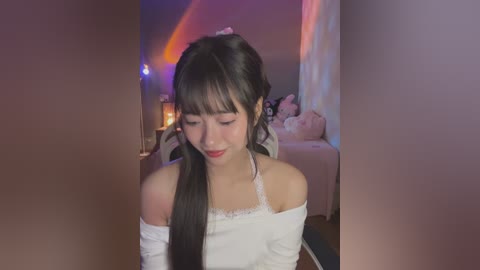 Media: Video of an East Asian woman with long black hair, wearing a white off-shoulder top, standing indoors with a cozy, dimly lit room in the background.