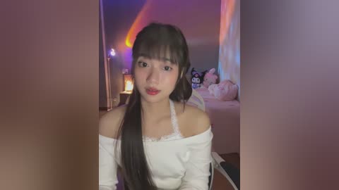 Media: Video of an East Asian woman with long, dark hair and fair skin, wearing a white off-shoulder dress, sitting in a dimly lit bedroom with neon lights and plush toys.