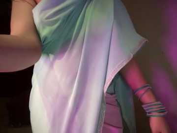 Media: A video of a person's arm and torso, draped in a light blue and pink shawl, with multiple colorful bangles on the wrist. The background is dark and indistinct.