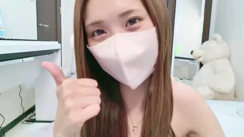 Media: A young Asian woman with long brown hair, fair skin, and wearing a white face mask and black top, gives a thumbs-up gesture in a modern, clean bathroom with white tiles and a plush teddy bear.