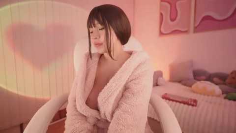 Media: A video of a young woman with a short bob haircut and fair skin, wearing a fluffy white robe, sitting in a white chair in a pastel-colored room with abstract art on the wall.