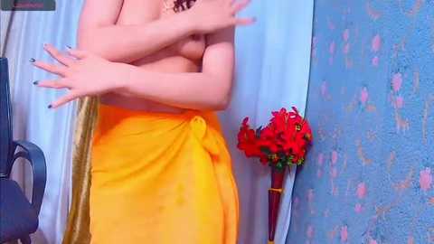 Media: Video of a topless woman with medium breasts, wearing a bright yellow sari, standing in front of a blue wall with red floral patterns.