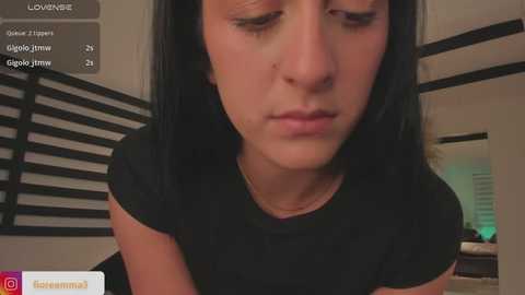 Media: Video of a young woman with long black hair, wearing a black top, looking down, in a modern, dimly-lit room with wooden slat wall and window blinds.