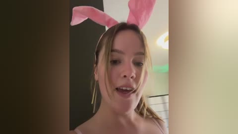 Media: Video of a young woman with fair skin, blonde hair, and pink bunny ears, wearing a pink top, smiling, indoors with a green wall and a ceiling light.