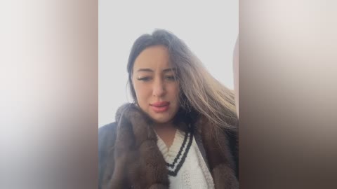 Media: Video of a young Latina woman with long, straight, dark hair, wearing a brown fur coat and white sweater, standing in a softly lit room with a blurred background.