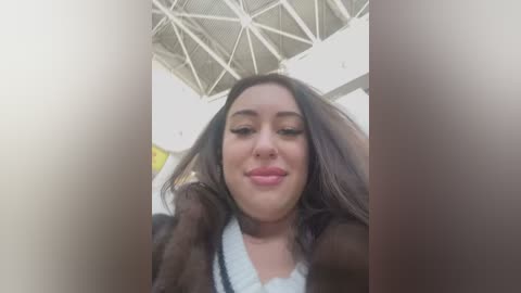 Media: Video of a smiling, light-skinned woman with dark hair and makeup, wearing a brown jacket and white top, standing in a modern, white building with a geometric ceiling.