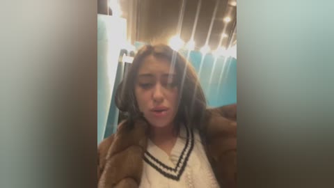 Media: A video of a young woman with medium-length, wavy brown hair, wearing a white lace top and a brown jacket, standing in a dimly lit, blue-tiled bathroom with string lights.